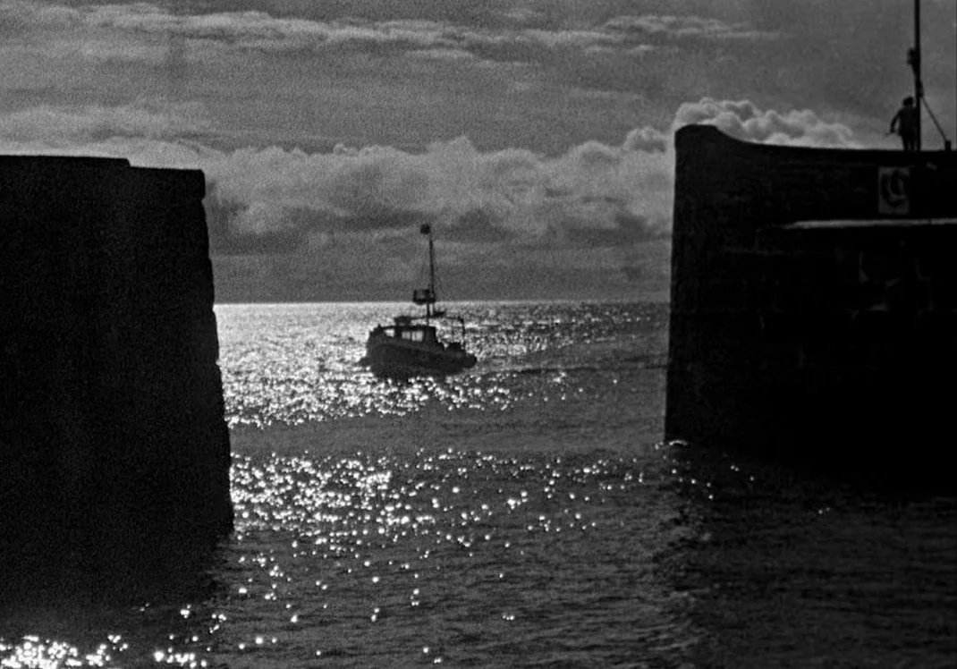 Silhouette of a boat sailing between two dark harbor walls on a sunlit sea under cloudy sky. A scene from the 2019 film Bait directed and written by Mark Jenkin, starring Edward Rower, Giles King, Mary Woodvine, Simon Shepherd.