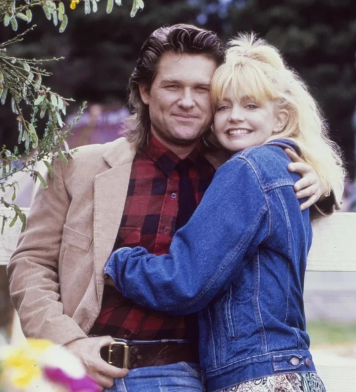 A film still from the rom-com film Overboard (1987) featuring Kurt Russell and Goldie Hawn. A man and woman looking happing and smiling hug each other.