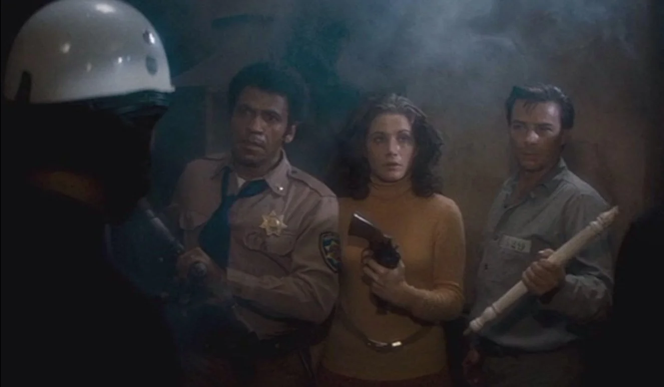 Three people in a dimly lit room with one woman holding a gun, and two men holding a pipe. This is a still from John Carpenters 1976 action film Assault on Precinct 13, starring Austin Stoker, Laurie Zimmer and Darwin Joston.