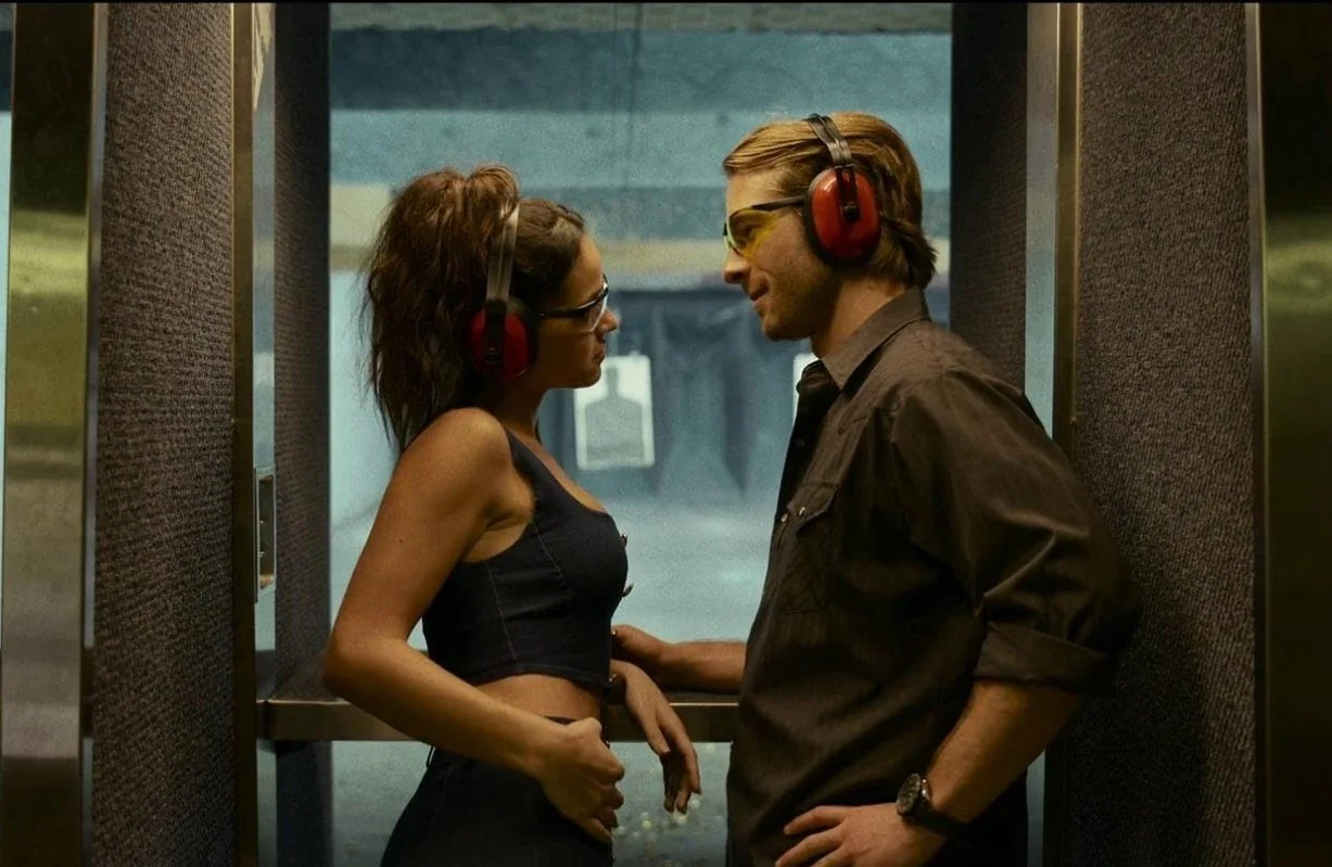 A still from the 2023 film Hit Man, starring Glen Powell and Adria Arjona. A man and a woman are standing in a shooting range facing each other, while wearing ear defenders and protective glasses.