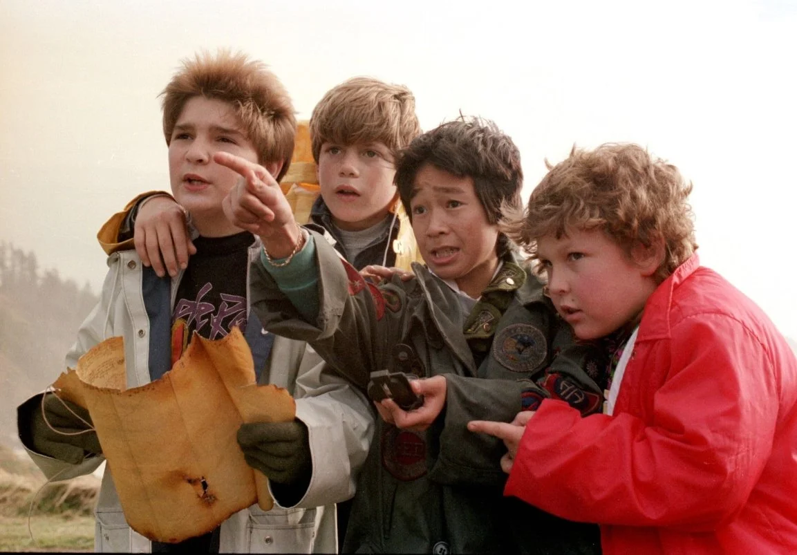 A film still from the Family adventure film The Goonies (1985) featuring Sean Astin, Corey Feldman, Ke Huy Quan and Jeff Cohen. A group of boys wearing rain coats are looking in the distance holding a map and a compass and pointing.