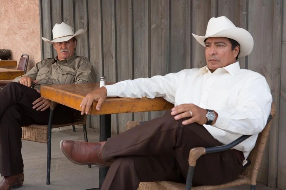 Two men wearing cowboy hats sitting at outdoor tables, one in a gray shirt and the other in a white shirt. This is a scene from the 2016 fil Hell or High Water starring Ben foster and Chris Pine.