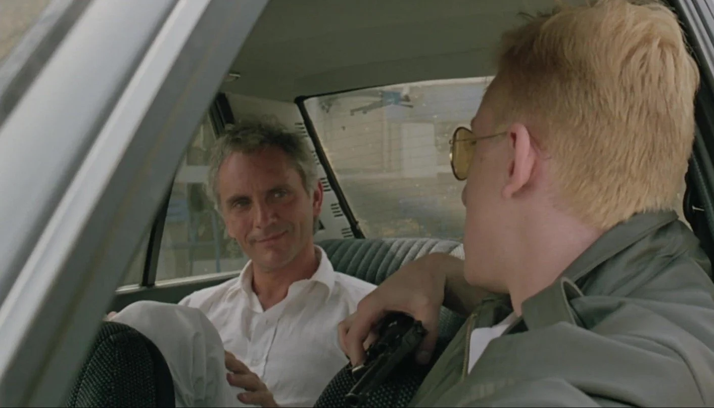 A film still from the crime gangster drama film The Hit (1984) featuring John Hurt and Tim Roth. Two men are sitting in a car, one in the front the other in the backseat, they are talking, one man has a gun.