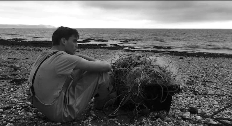 A person sitting on a rocky beach with a tangle of fishing ropes and gear, overlooking the ocean under an overcast sky. From the 2019 film Bait directed by Mark Jenkin starring Edward Rowe and Isaac Woodvine.