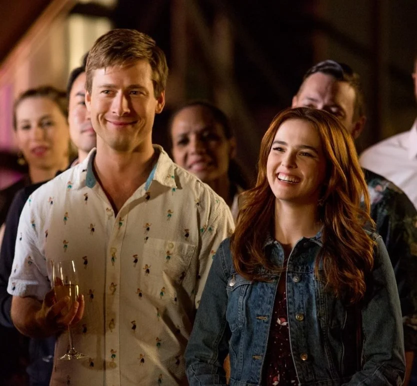 A film still from the rom-com film, Set It Up (2018) featuring Glen Powell and Zoey Deutch. A man and woman are standing next to each other smiling.