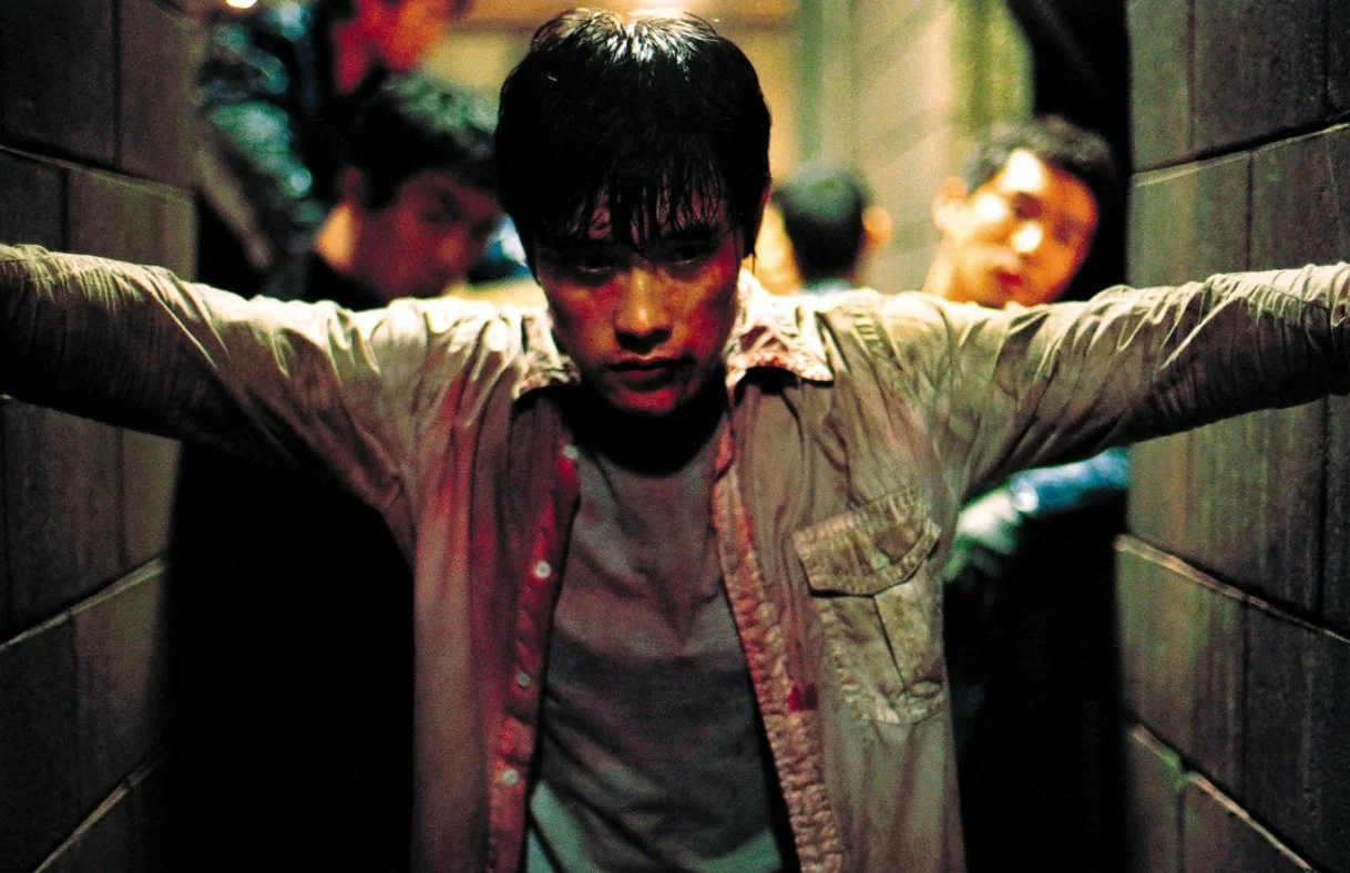 A man wearing a white shirt covered in blood with his arms outstretched. Three other young men are in the background in a dimly lit, narrow corridor. This is a still form Kim Jee-woon's 2005 film A Bittersweet life starring Lee Byung-hun.