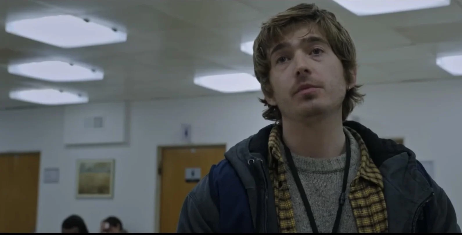 A young man with shaggy brown hair, wearing a jumper layered with a plaid shirt and a dark jacket. He is in an in a hospital with fluorescent ceiling lights. Resident Evil (2026) directed by Zach Cregger and starring Austin Abrams.