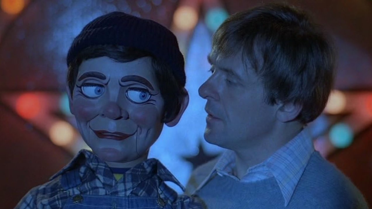 A man looking at a ventriloquist dummy with exaggerated facial features in a dimly lit setting with colorful blurred lights in the background. Scene from the 1978 film Magic Directed by Richard Attenborough starring Anthony Hopkins
