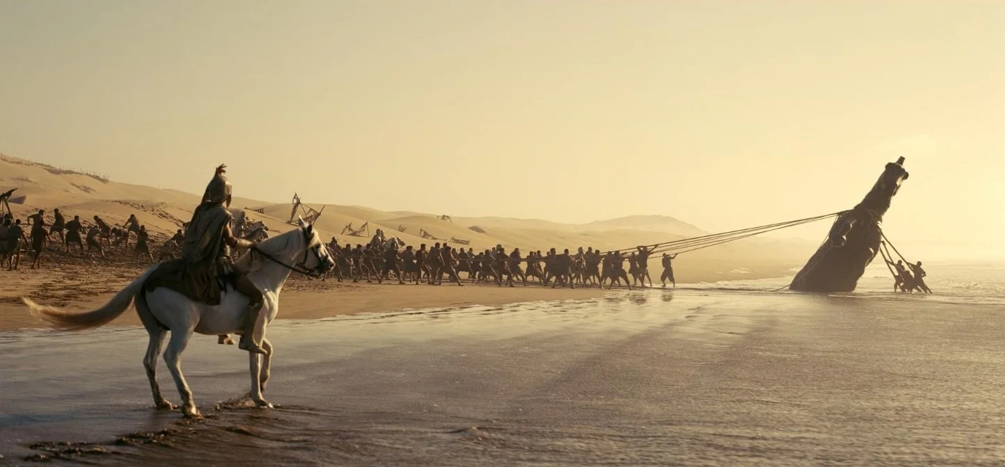A large group of people riding horses along the shoreline in a desert landscape, they are pulling a submerged wooden horse from the water. This is a still from the film The Odyssey (2026) directed by Christopher Nolan and stars Matt Damon.