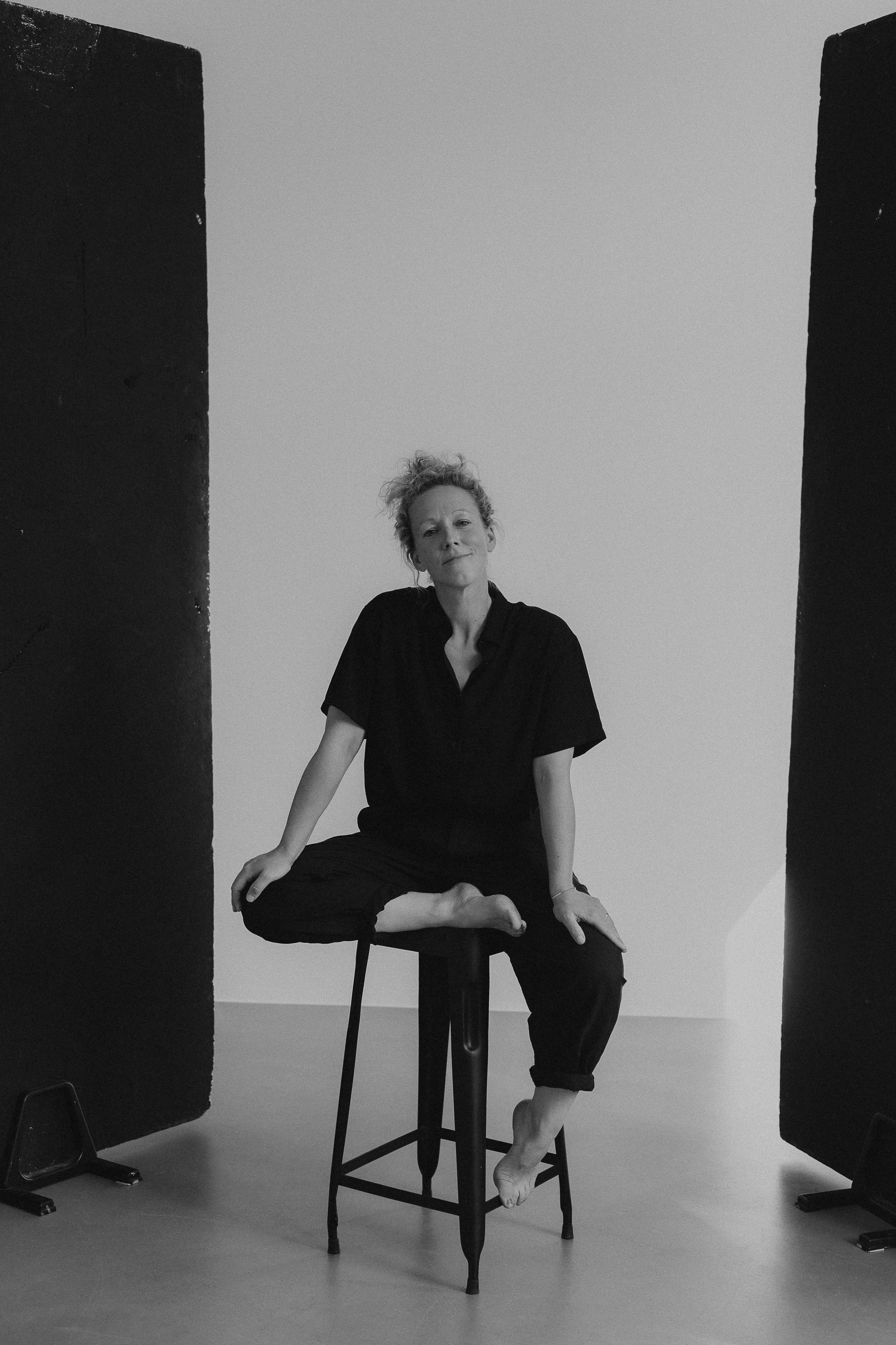 Schwarz-weiß-Fotografie einer Frau, die barfuß auf einem Stuhl sitzt, umgeben von zwei schwarzen Trennwänden, eines mit einem Stuhl, das andere ohne. Sie trägt eine dunkle Bluse und schwarze Hosen, sitzt in einer entspannen Haltung, mit leicht hochgelegtem Haar und einem leichten Lächeln.