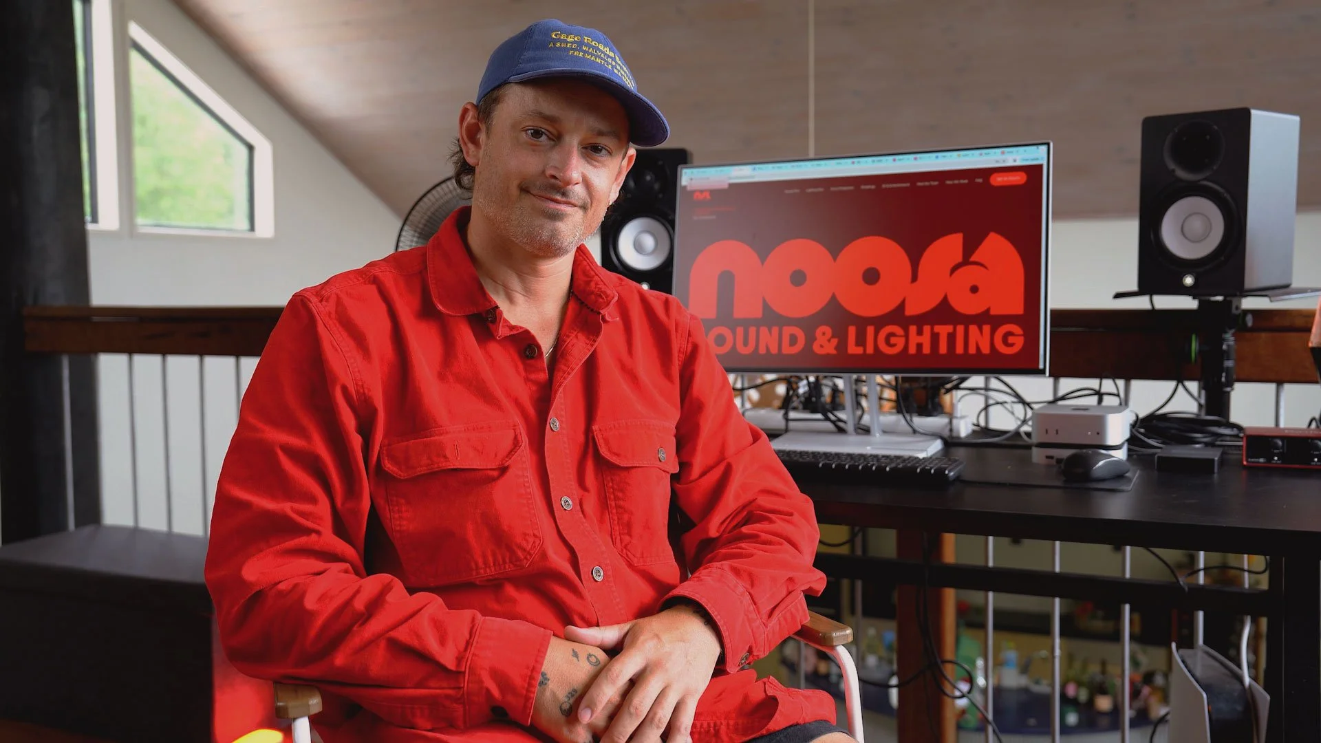A man in a red shirt and blue baseball cap sitting in a room with a computer setup, including speakers and a monitor displaying the words 'Noosa Sound & Lighting.'