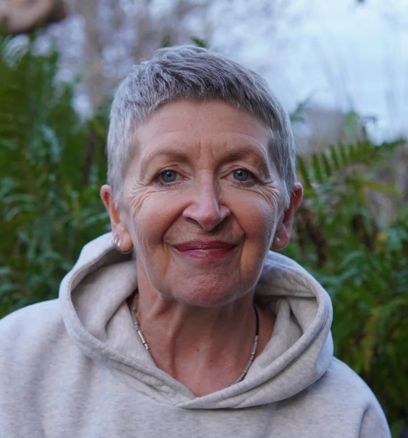 Close-up of an elderly woman with short gray hair, smiling, wearing a light-colored hoodie, outdoors with green foliage in the background.