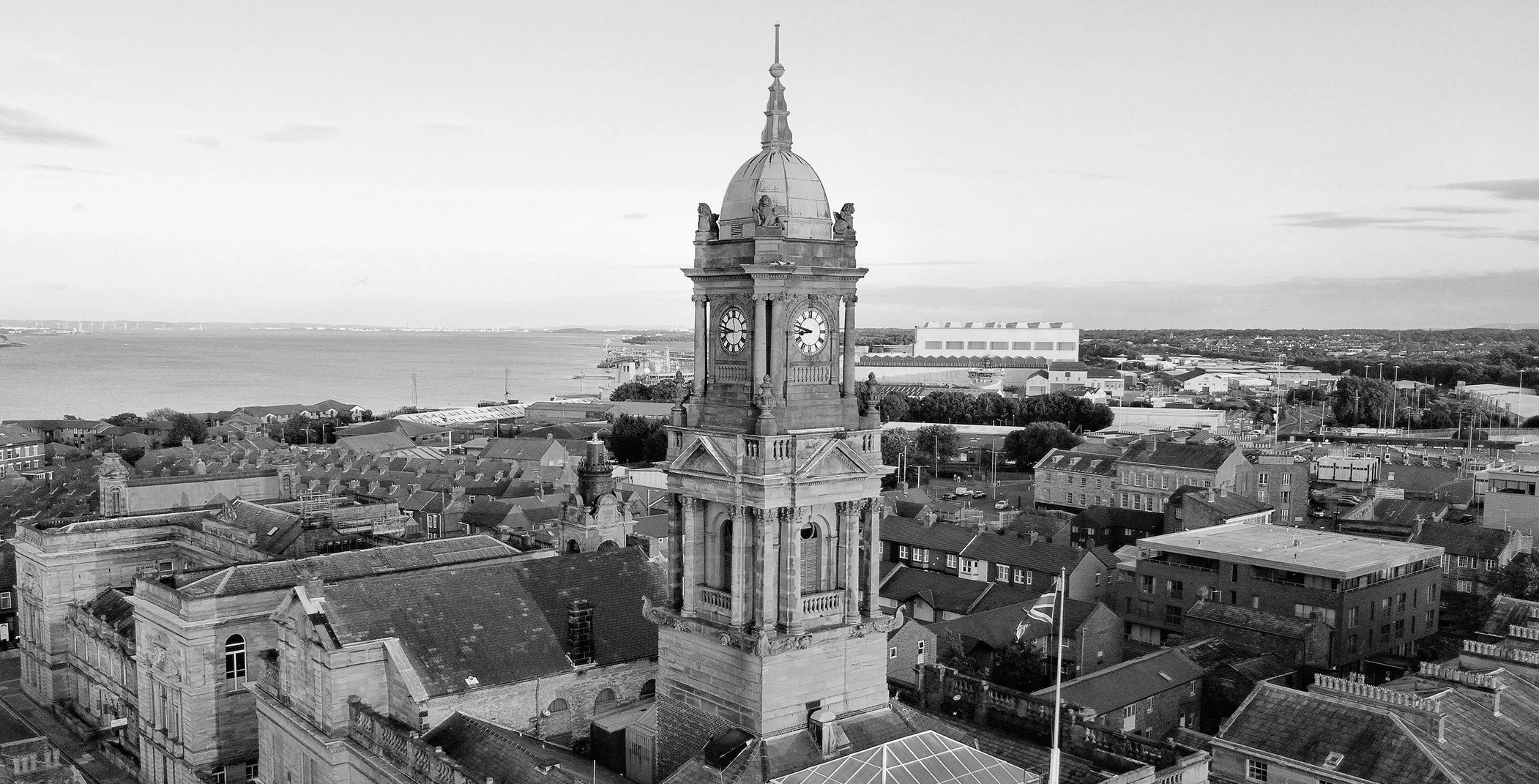 Black and white aerial view of a town with a prominent clock tower, water in the background, and industrial buildings nearby.