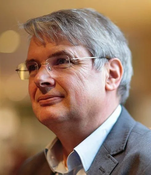 Close-up van een man met grijs haar en bril, gekleed in een grijze jas en witte overhemd, lachend en kijkend naar links.