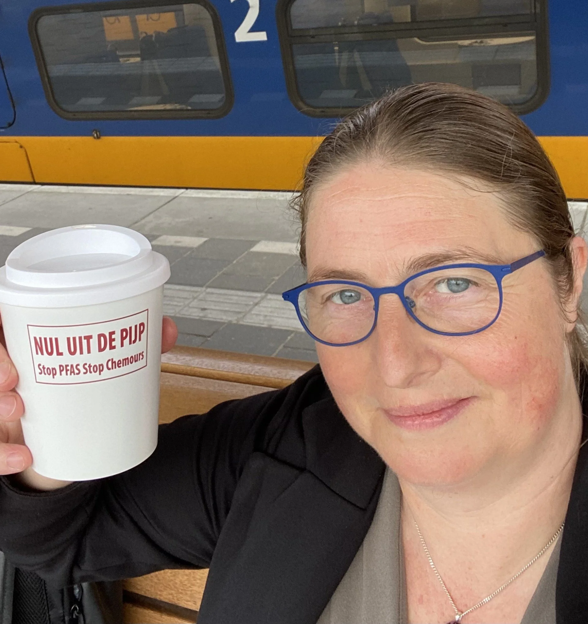 Een persoon met blauwe brillen die een kopje koffie vasthoudt met de tekst 'NUL UIT DE PUP Stop PFAS Stop Chemours' erop, zittend op een bankje op een treinhalte.