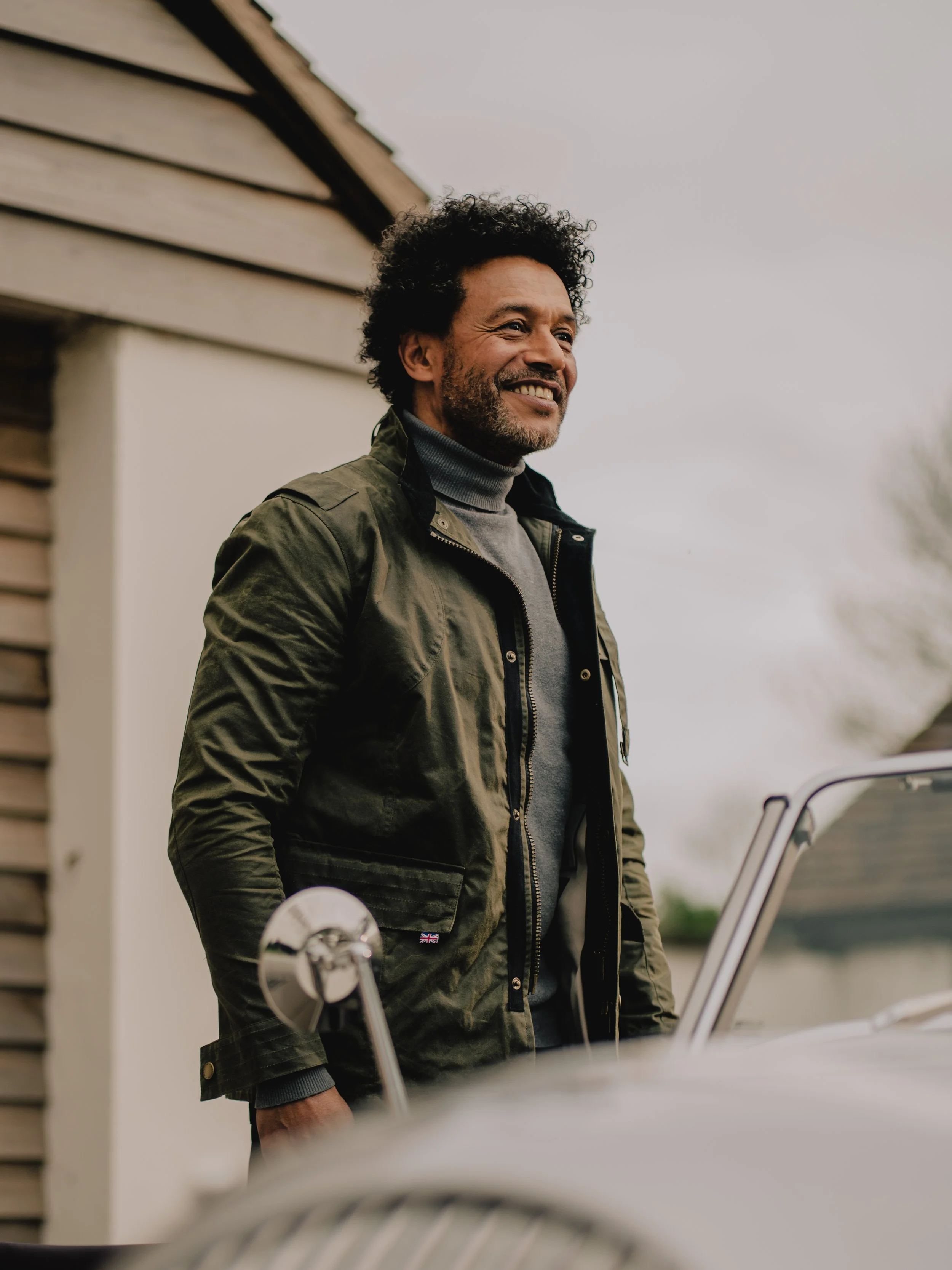 Smiling man with curly hair and beard standing outside near a house, wearing a green jacket and gray turtleneck, with a car in the foreground.