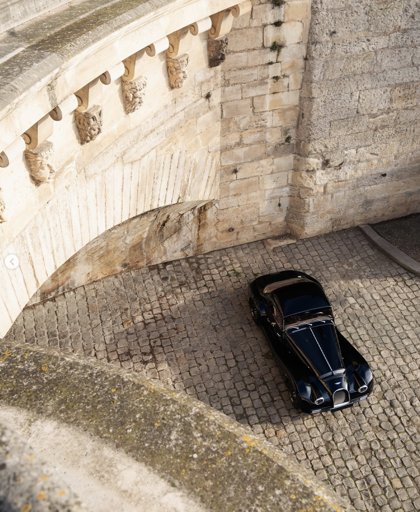 View of a cobblestone courtyard with a black luxury car parked near an old stone wall with decorative architectural details.