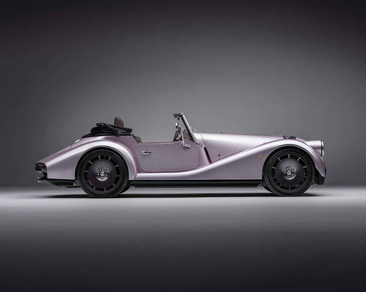 A sleek, silver, vintage convertible sports car with black wheels, parked in a studio with a gray background.
