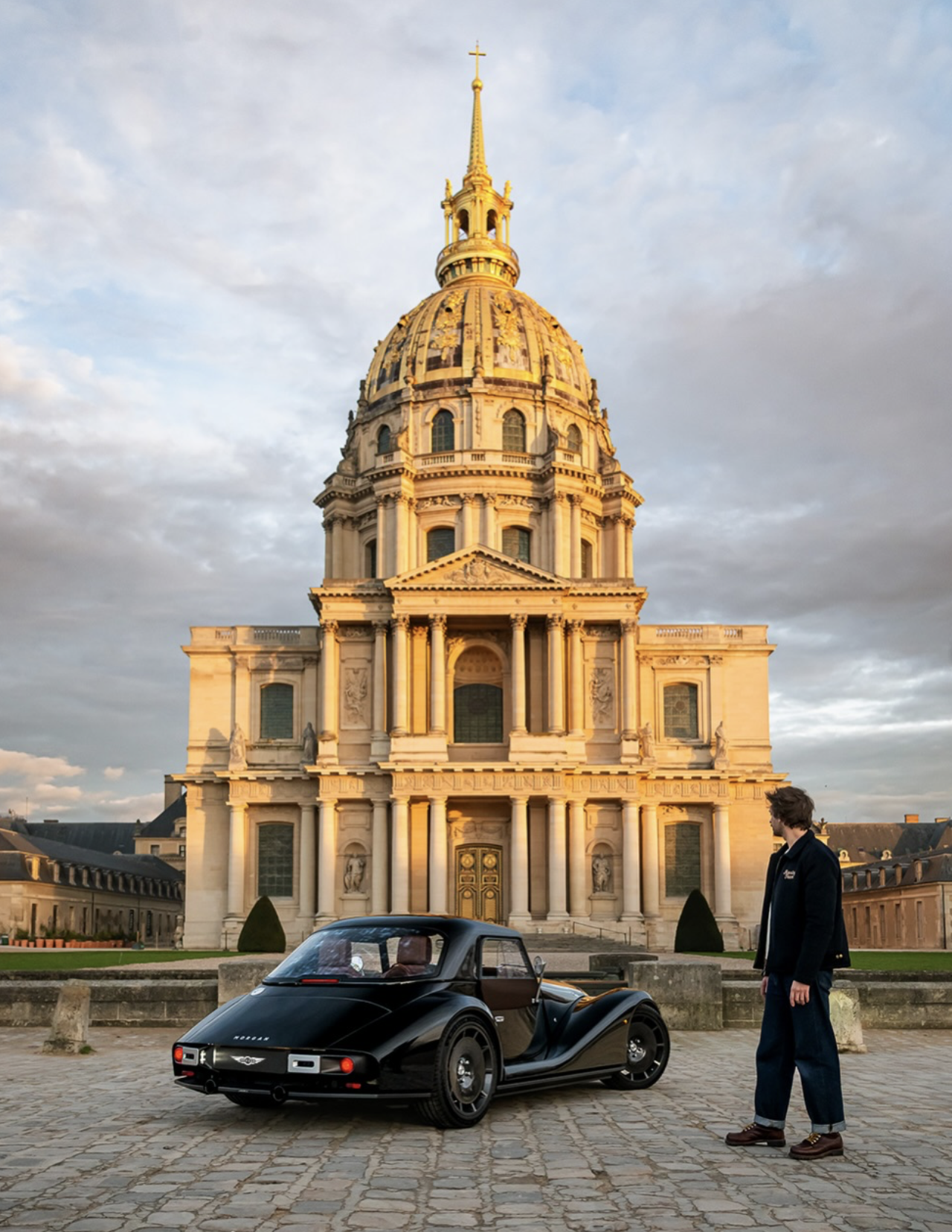 A man standing next to a vintage black mini car in front of a historic gold-domed building, possibly during sunset with a partly cloudy sky.