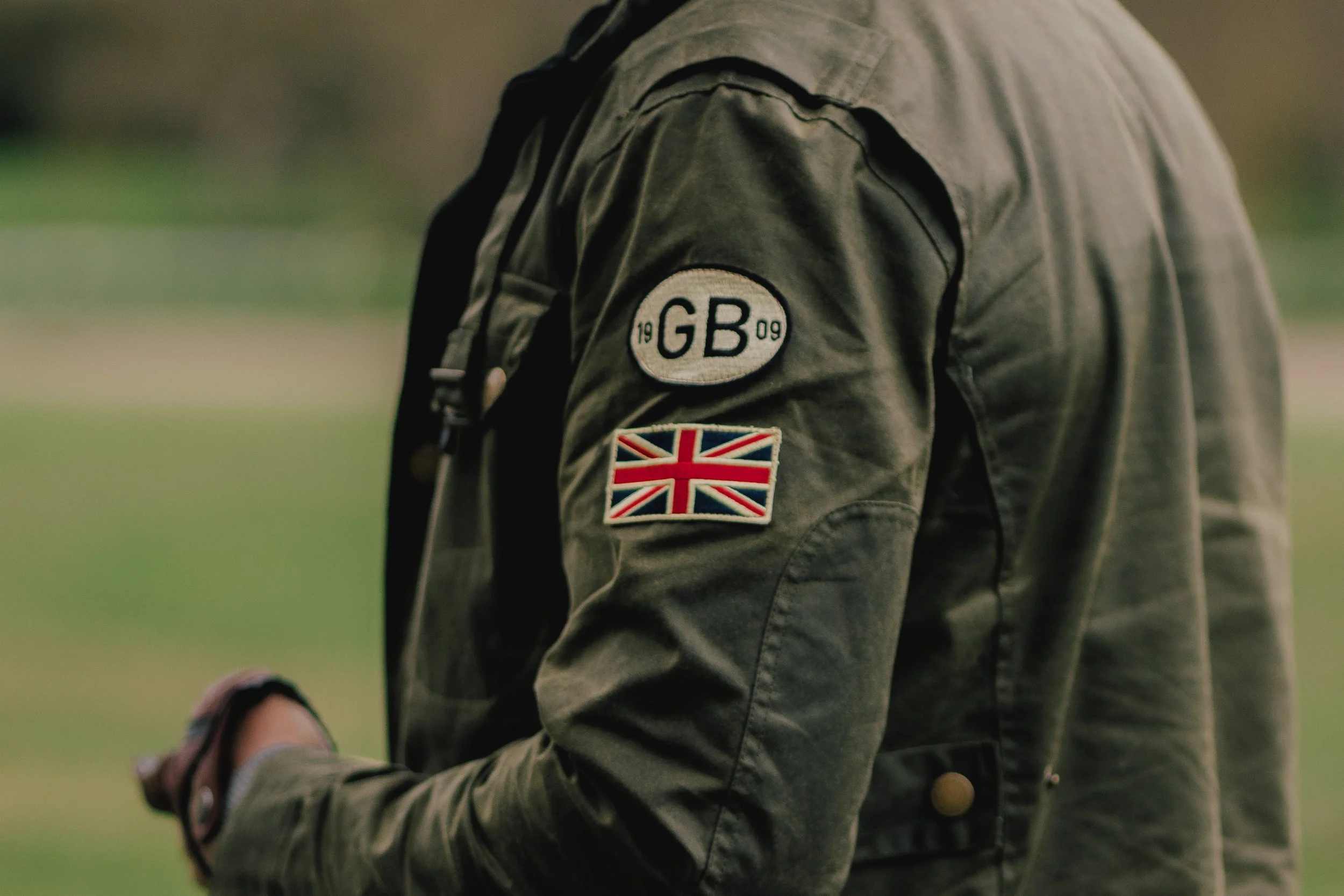 Close-up of a person wearing a military-style jacket with a patch reading "GB 1909" and a British flag patch, holding a sports ball.