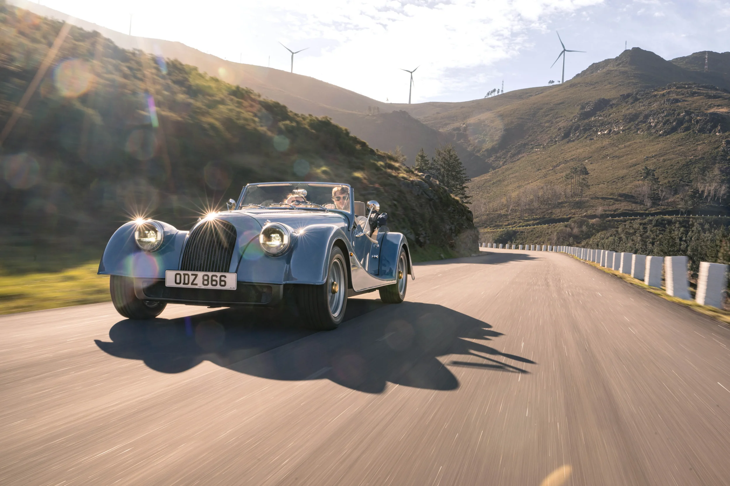 A vintage blue convertible car with two people inside, driving on a curved mountain road with wind turbines and hills in the background during daytime.