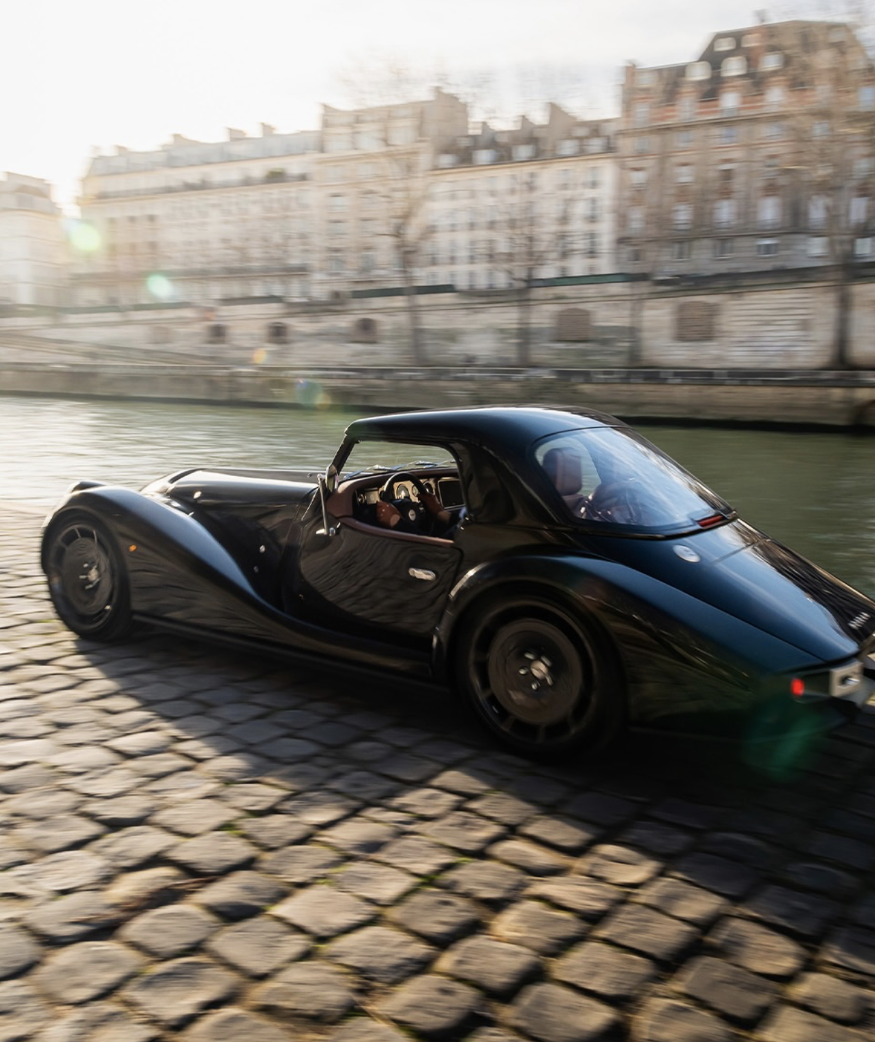A black vintage-style car driving along a cobblestone riverbank with Parisian buildings in the background.