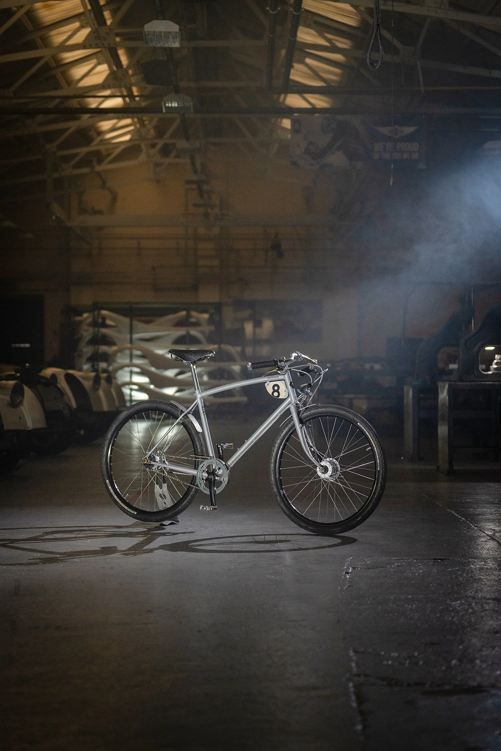 A silver mountain bike with a black saddle, parked inside a dimly lit indoor garage with vintage cars and a motorcycle in the background.