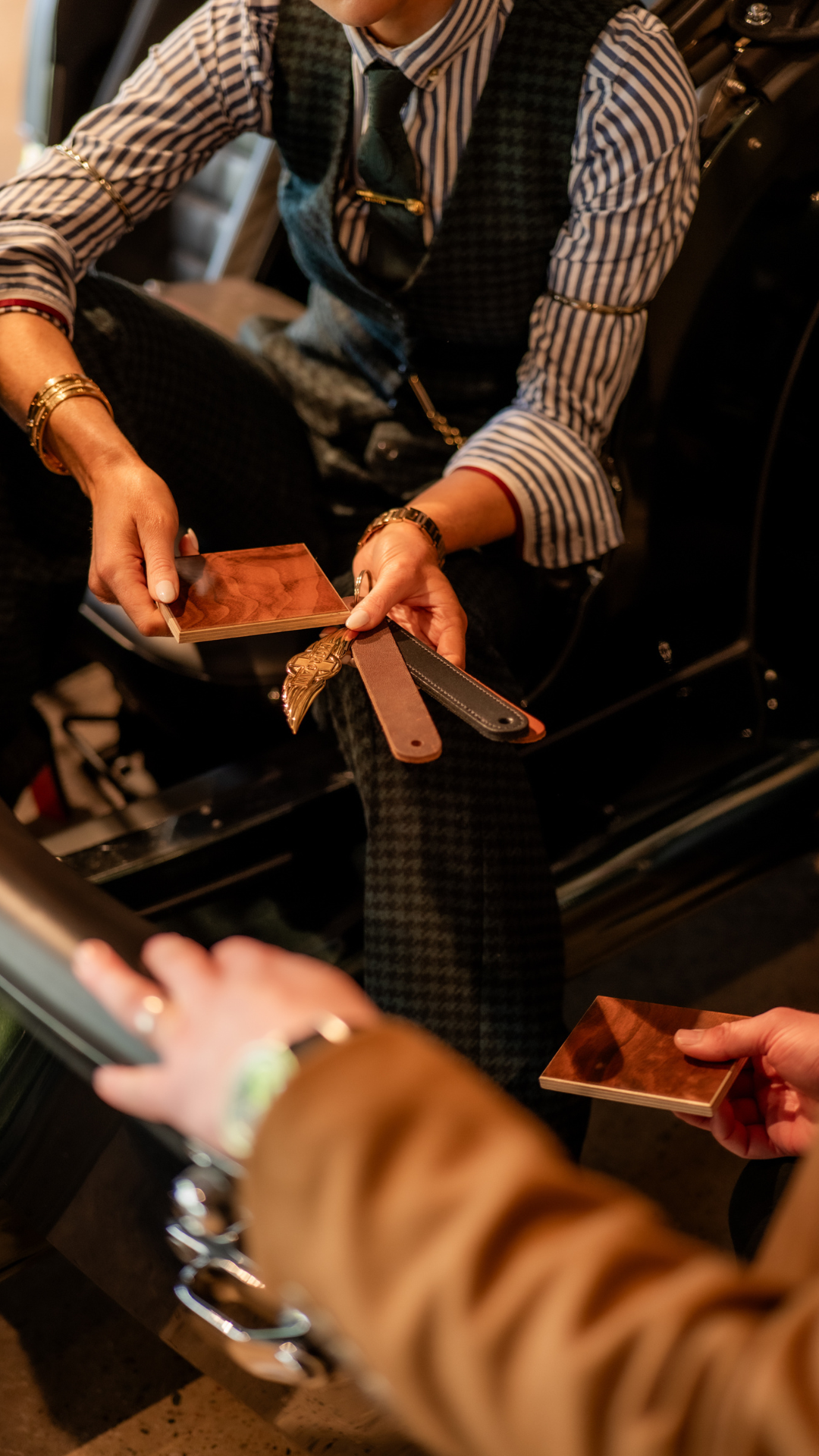 Person in a striped shirt and vest exchanging small wooden color sample cards with another person, holding various leather sample strips in their hand.