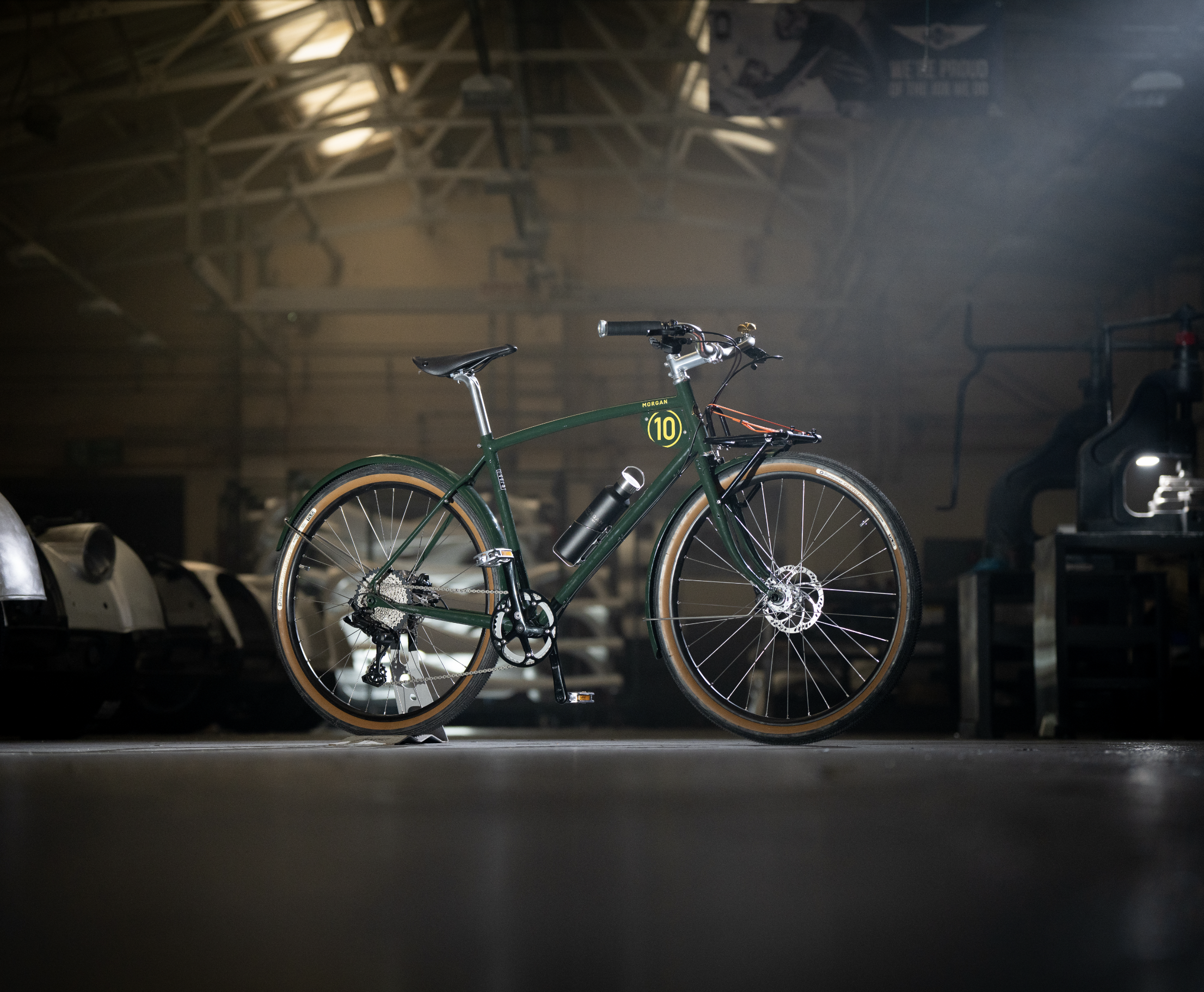 A green mountain bike with brown tires and a black water bottle mounted on the frame in a dimly lit garage or warehouse with exposed wooden ceiling beams.