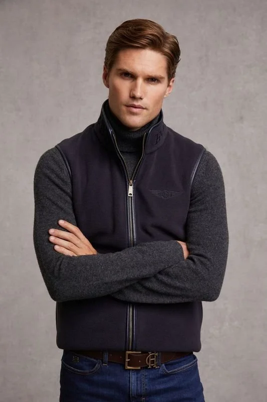 A young man with brown hair and a serious expression, wearing a dark gray turtleneck, a navy vest, and blue jeans, standing with arms crossed against a plain gray background.