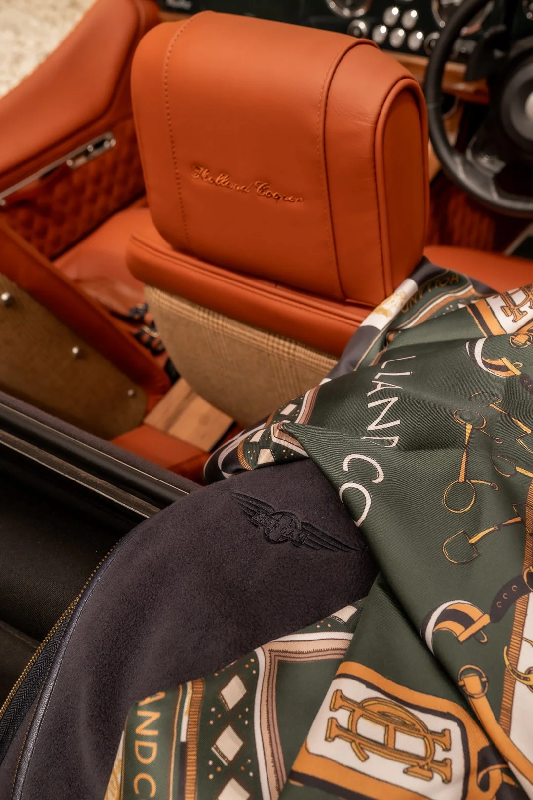 Inside a car showing a tan leather bag with 'The Blind Tiger' embossed on it, a dark cloth with a winged emblem, and a colorful scarf with gold, black, white, and green patterns draped over a person's leg.