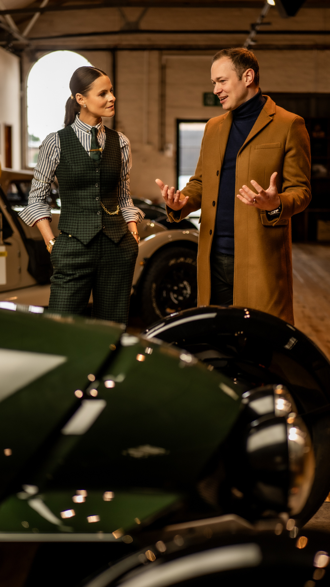 A man and woman are talking inside a garage filled with classic sports cars, including a green one in the foreground and a white one behind them. The man is dressed in a brown coat and navy turtleneck; the woman is wearing a checkered vest, striped shirt, and accessories, with her hands in her pockets.