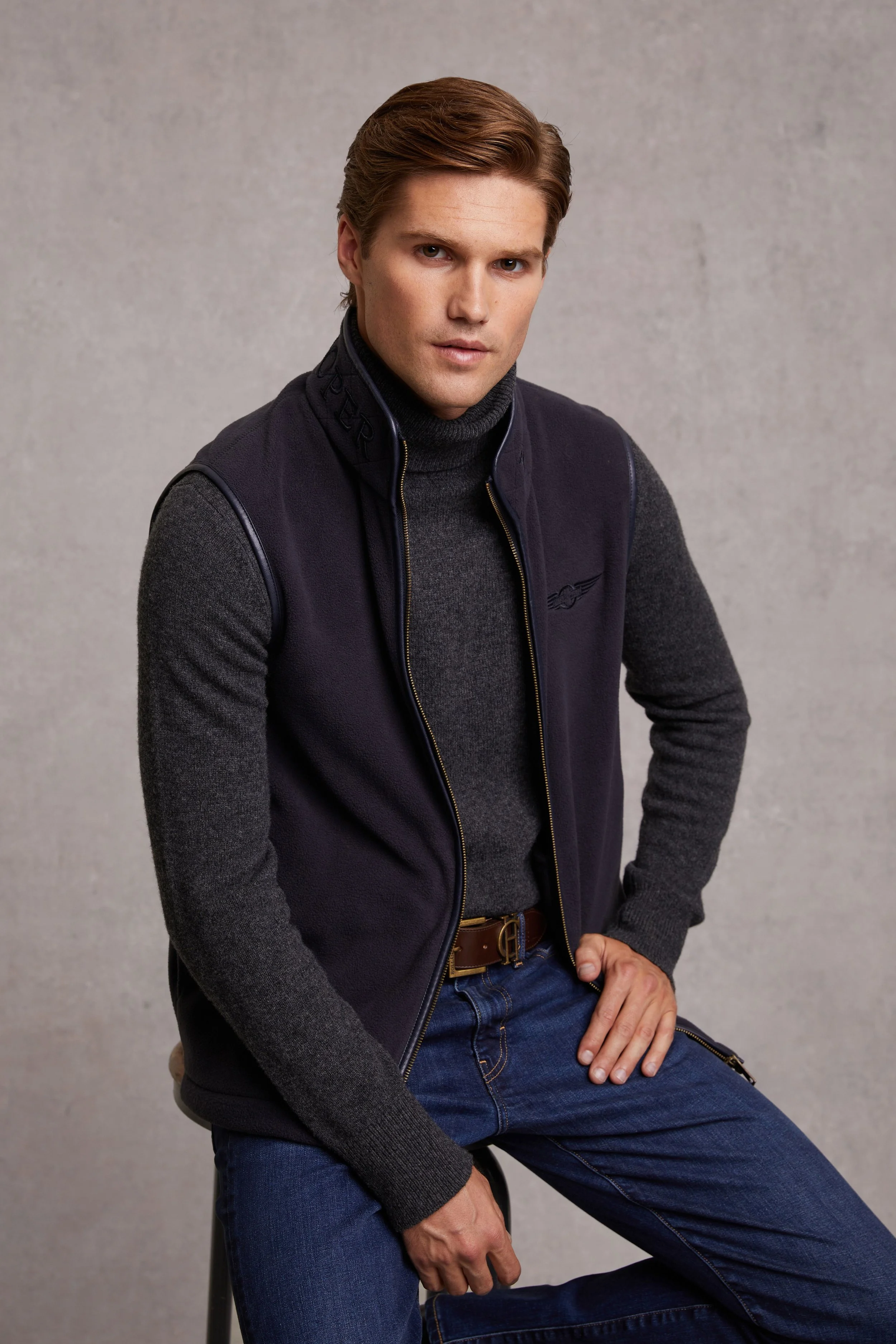 A young man with brown hair wearing a dark vest over a gray turtleneck sweater and blue jeans, sitting against a plain gray background.