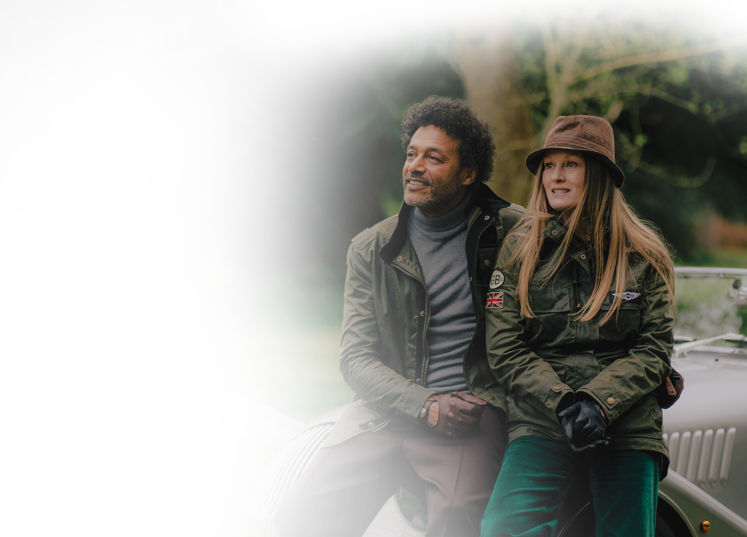 A man and woman sitting on a park bench in a green park with trees. The man has curly hair and is wearing a jacket, and the woman has long hair, a hat, and a jacket. They are smiling and looking off into the distance.