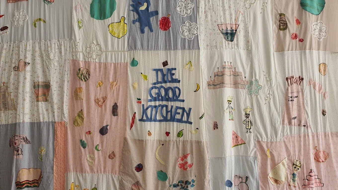 A patchwork quilt with various embroidered and painted designs, including fruits, vegetables, a birthday cake, and the phrase "THE GOOD KITCHEN" in blue letters.