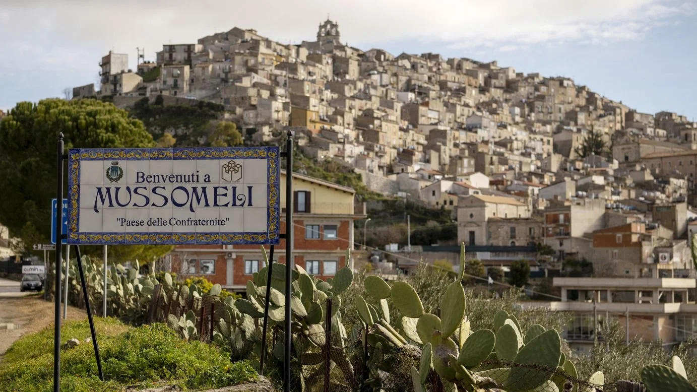 Sign welcoming visitors to Mussomeli, Italy, with a hillside town in the background and cactus plants in the foreground.
