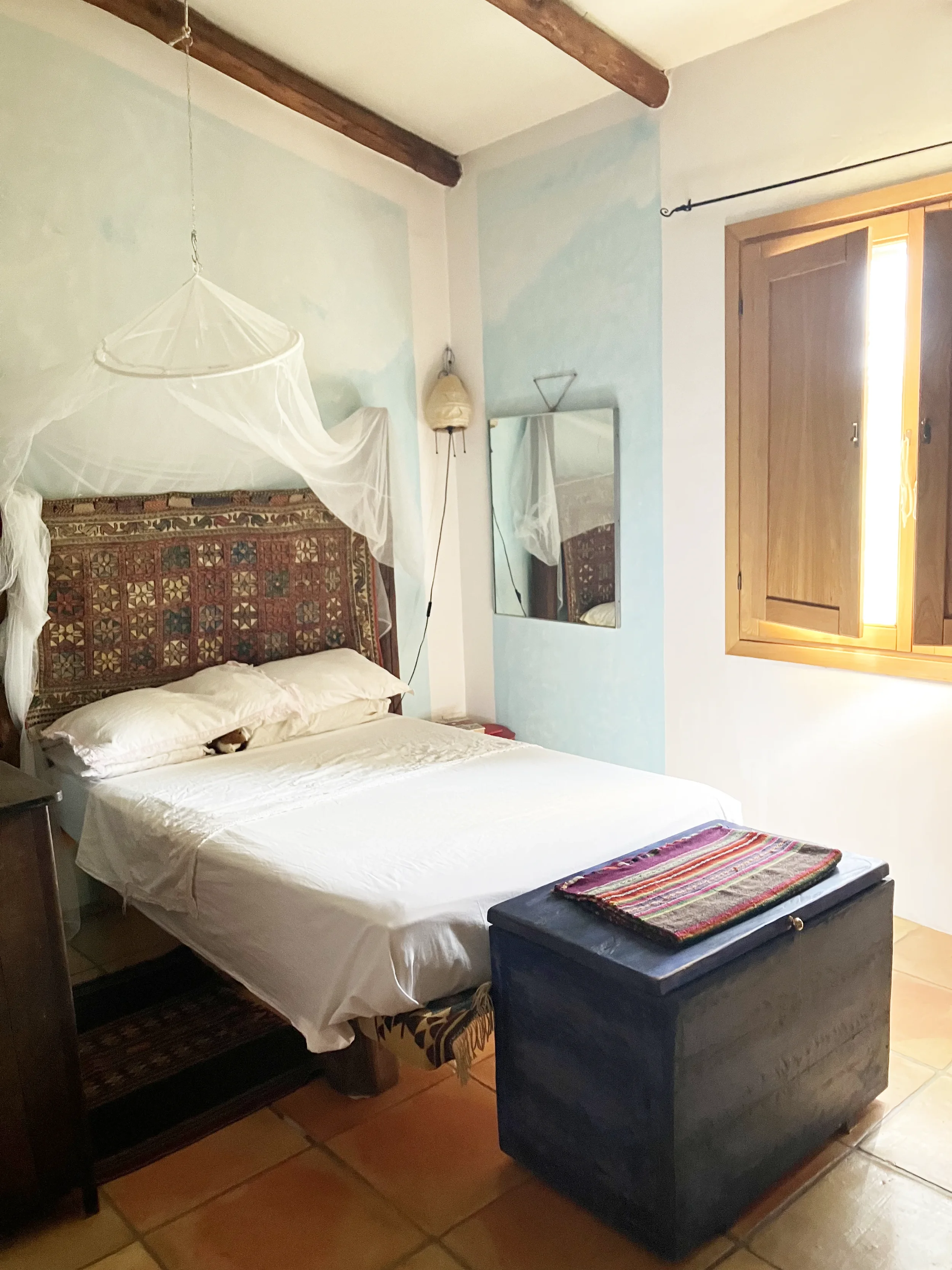 Ein Schlafzimmer mit einem Bett, das mit weißen Bettwäsche und Kissen bedeckt ist. Es gibt ein Stoff-Pendeltuch über dem Kopfteil des Bettes, einen Holzfenster mit geschlossenen Jalousien und einen Spiegel an der Wand.