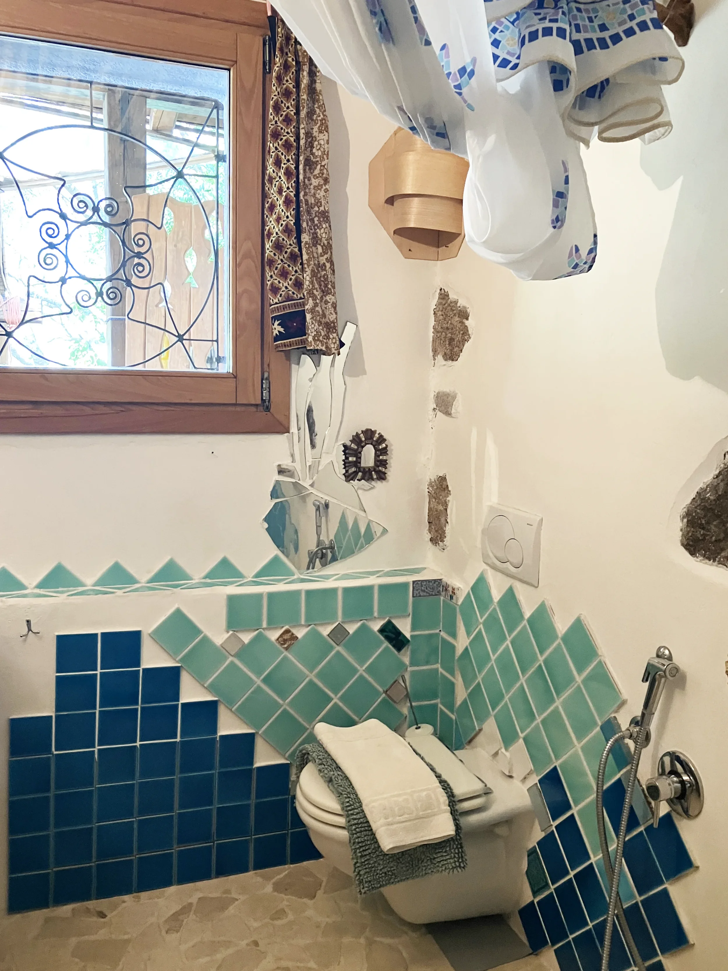 Ein Badezimmer mit einem Fenster, türkischen Fliesen, einem geöffneten WC mit Handtuch, einem Spiegel, einer Wasserarmatur und einer Duschbrause. Es ist teilweise renoviert, einige Stellen an der Wand sind offen.