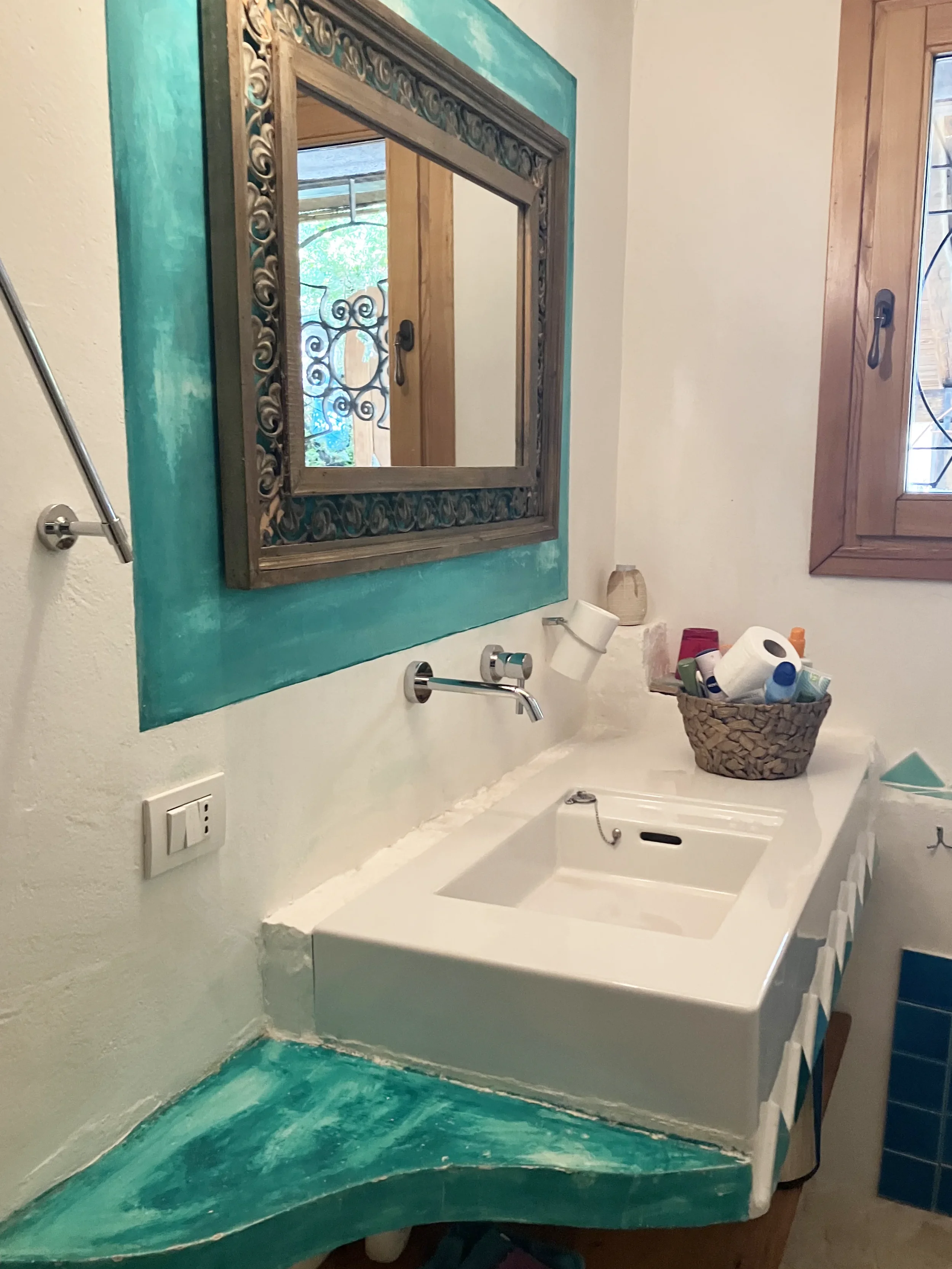 Badezimmer mit weißem Waschbecken, Wandspiegel und zwei Fenster, eines mit Holzrahmen und Metallgitter. Es gibt eine Waschschüssel mit Badezimmerartikeln, ein Handtuch und eine Steckdose an der Wand.