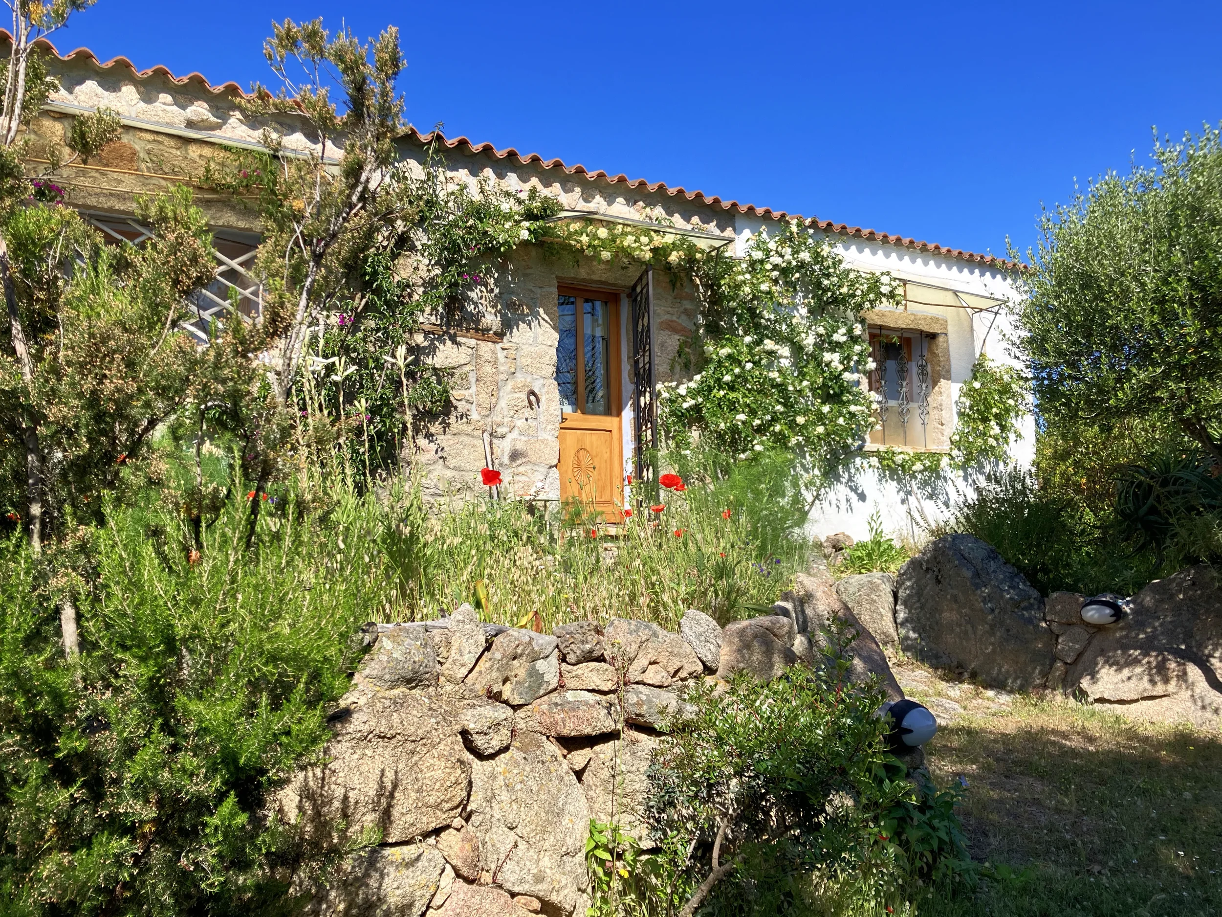 Ein kleines Haus mit Stein- und weißen Wänden, umgeben von Garten mit Blumen und Bäumen, unter blauem Himmel.