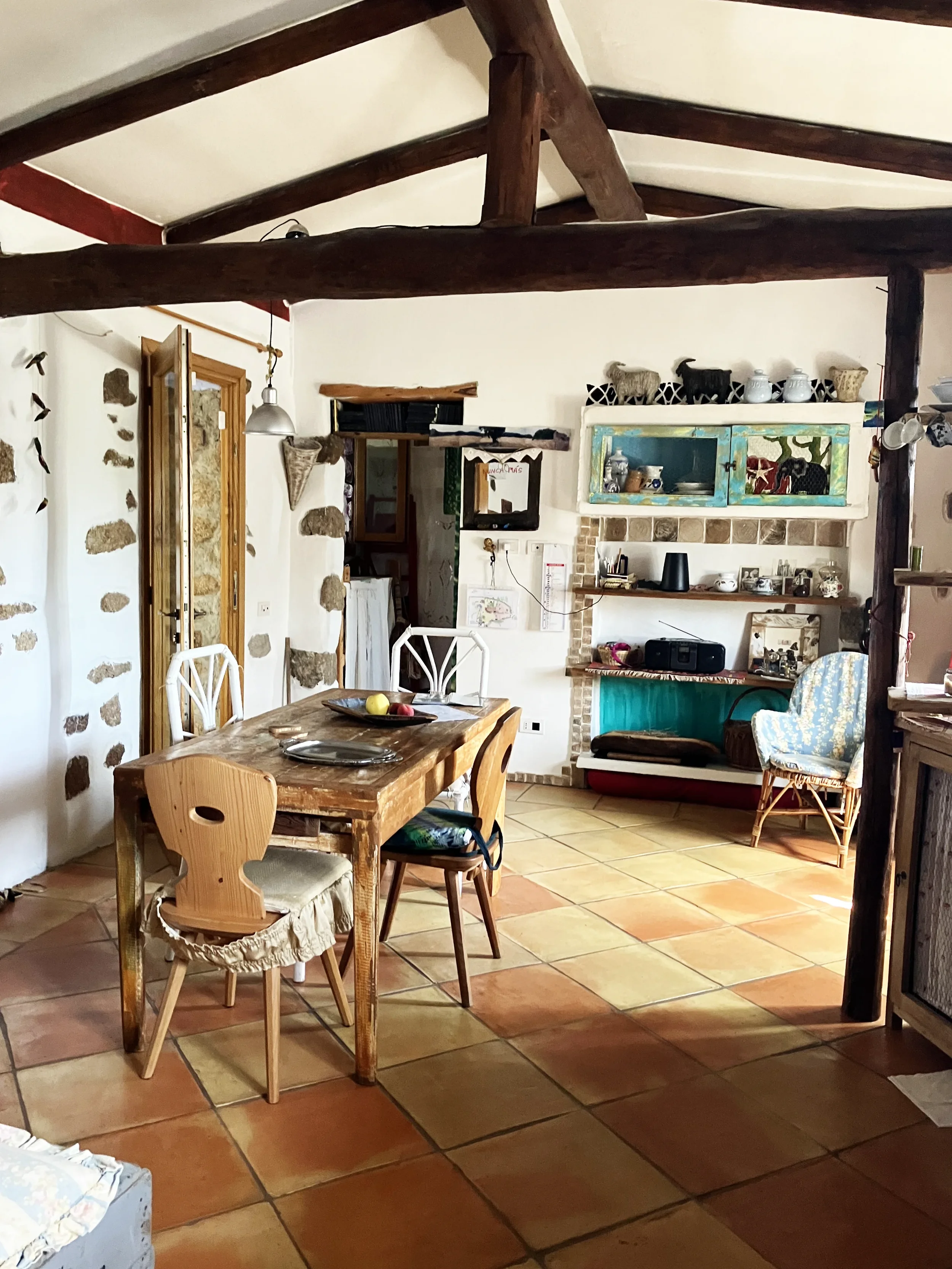 Innenansicht eines rustikalen Esszimmers mit Holzdeckenbalken, einem Holztisch mit vier Stühlen, einer Wand mit Steinen und einer offenen Küche im Hintergrund.