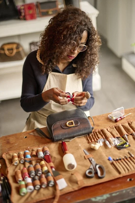 A woman with curly hair and glasses sits at a wooden table, working on a sewing project. She has a beige apron on and is surrounded by spools of thread, scissors, and other sewing tools. There is a black purse on the table and a small plastic container with sewing supplies.