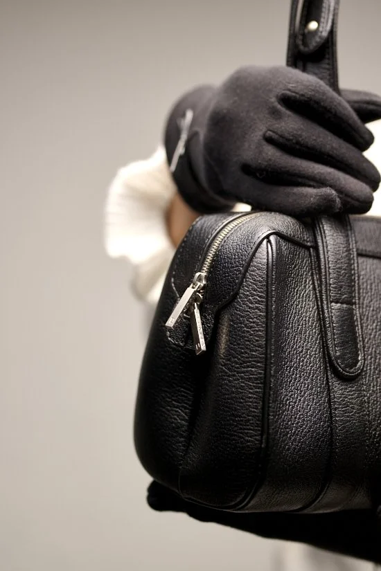 Close-up of a person holding a black textured leather handbag, wearing black gloves and a white face mask.