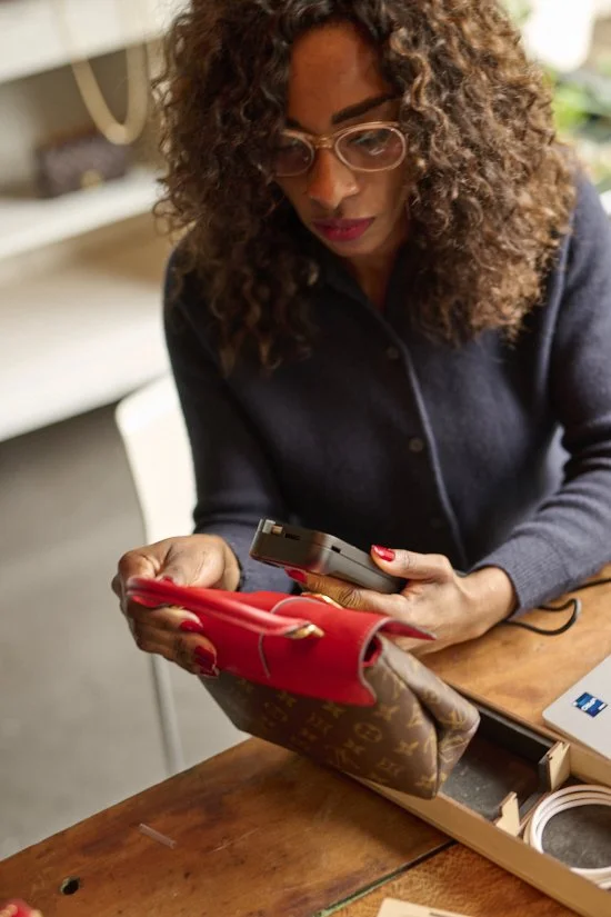 A woman with curly hair, glasses, and red-painted nails, is sitting at a wooden table, looking at her phone inside a store or café. There is a Louis Vuitton bag and an opened box with a charging cable on the table.