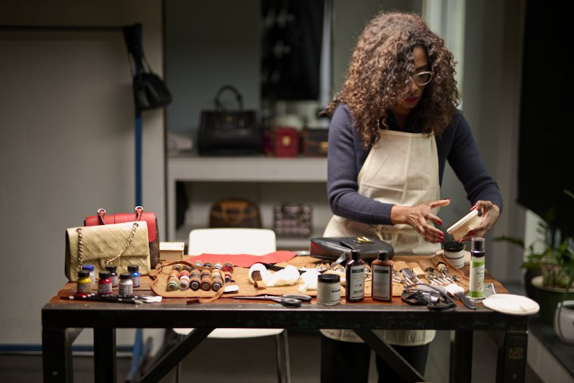 A woman with curly hair and glasses wearing an apron stands at a table, organizing leather crafting supplies including thread spools, dye bottles, and tools in a workspace.