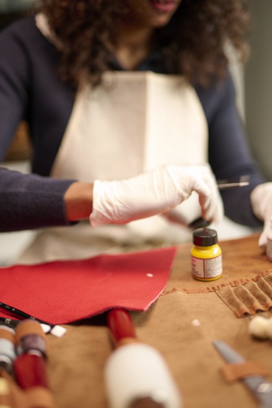 Person wearing gloves and apron working on a craft project with small bottles and tools on a table.