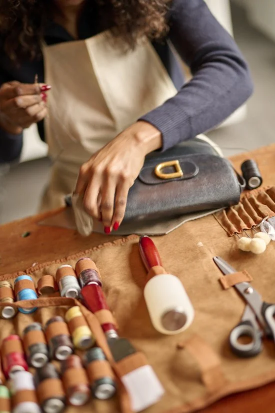 Close-up of a woman sewing on a black leather purse with a gold buckle, surrounded by sewing supplies including spools of thread, scissors, and a sewing roller, on a brown fabric-covered table.