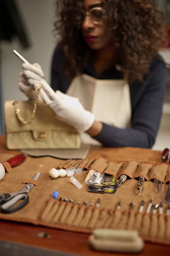 A woman with curly hair wearing gloves and a beige apron, working on jewelry with tools and supplies laid out on a wooden table.