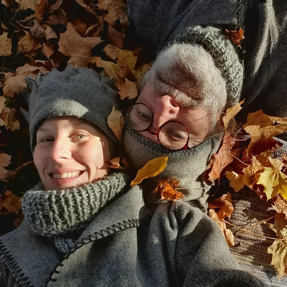 Två personer ligger på höstlöv, en kvinna och en man, båda nära varandra, bärandes mössor och varma kläder, solsken i ansiktet.