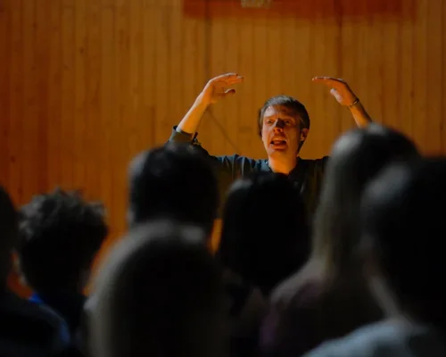 En person håller händerna i luften och talar till en publik i ett rum med trägolvsväggar.